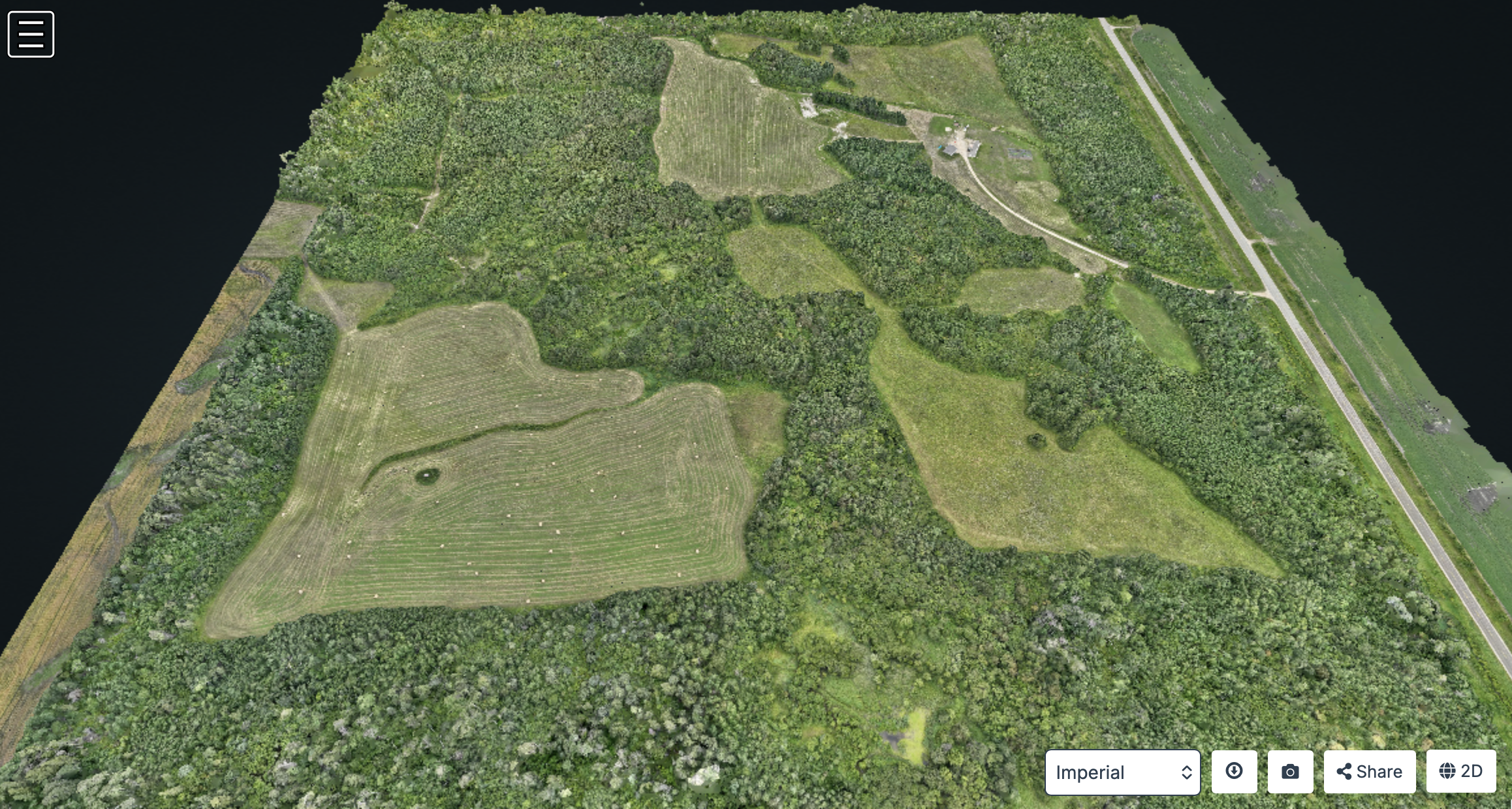 Aerial view of a large green landscape with trees, grassy fields, and a water body on the left side. There is a small structure and a winding road on the right side.