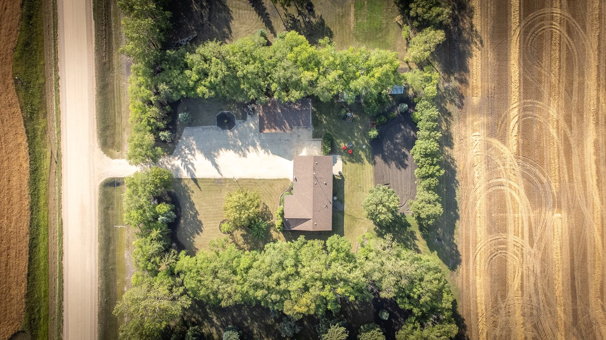 An aerial view of a house surrounded by trees, two sheds, a yard, and neighboring fields with a dirt road on the left side real estate photography Manitoba.