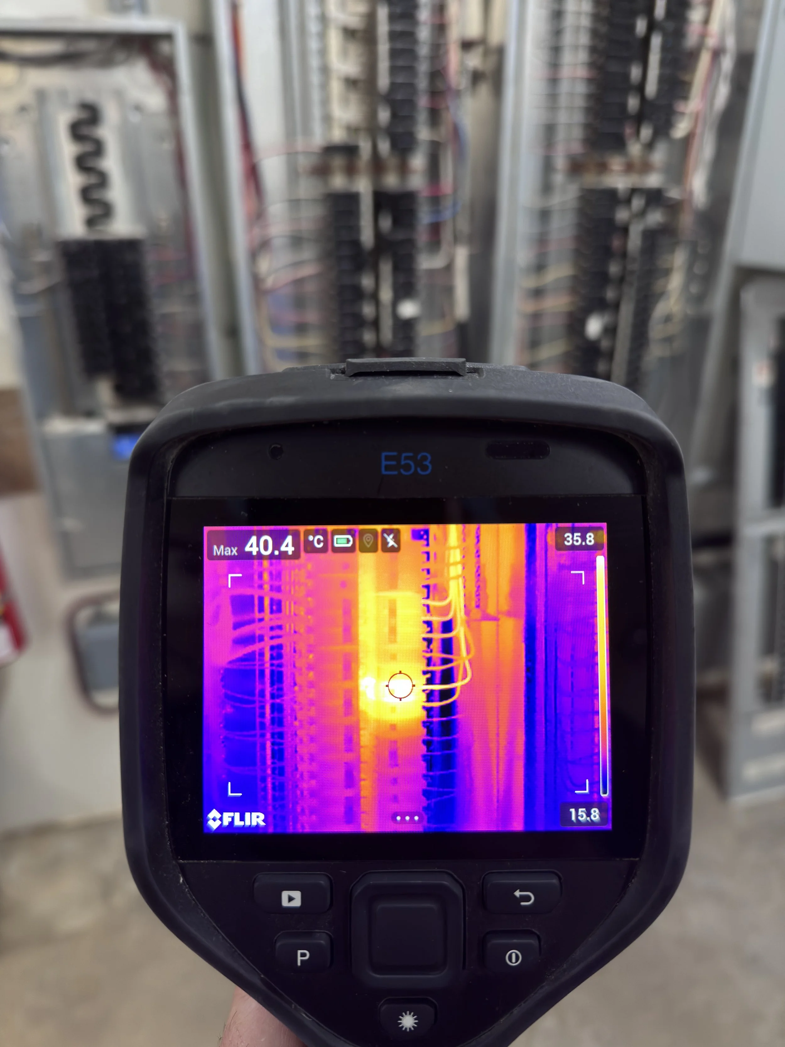 A thermal imaging camera displays an image of electrical panels or circuit breakers, highlighting heat variations with bright colors indicating higher temperatures.