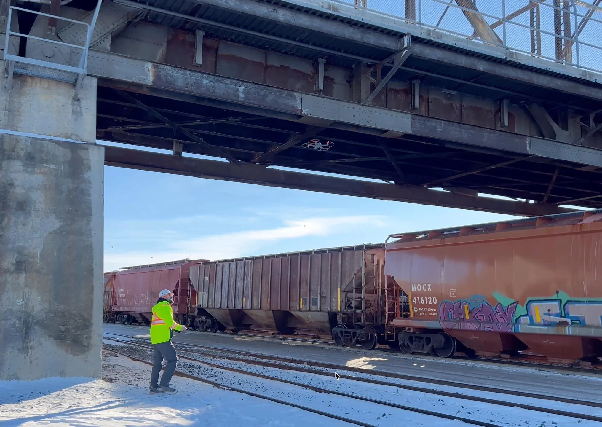 Rail yard worker in a high-visibility jacket beside graffiti-covered freight cars beneath a steel bridge. manitoba bridge and infrastructure  inspections service salter bridge