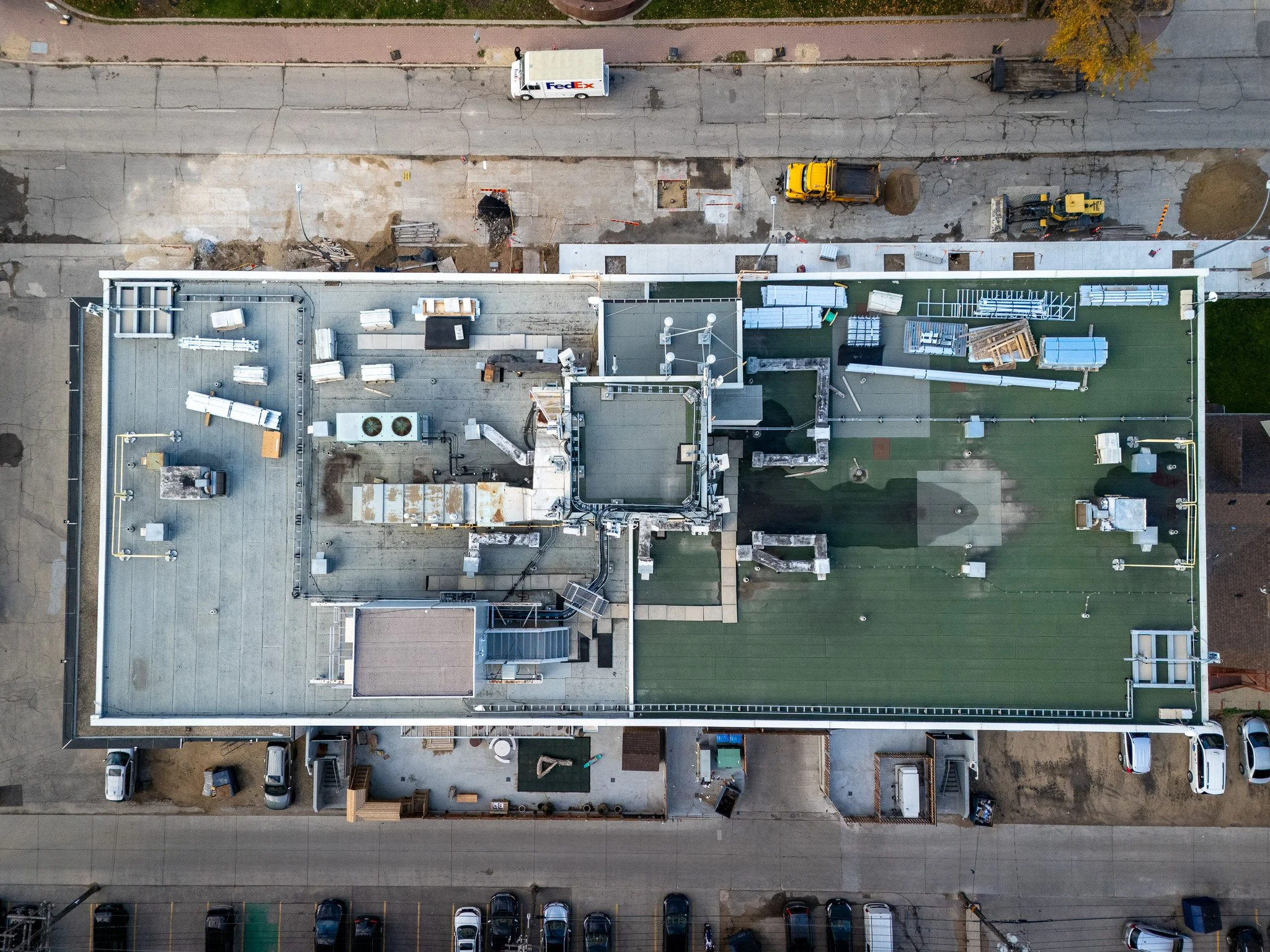 An aerial view of a rooftop under construction, with construction equipment, materials, and vehicles, next to a cracked sidewalk and a FedEx delivery truck parked above. aerial roof inspection  Winnipeg