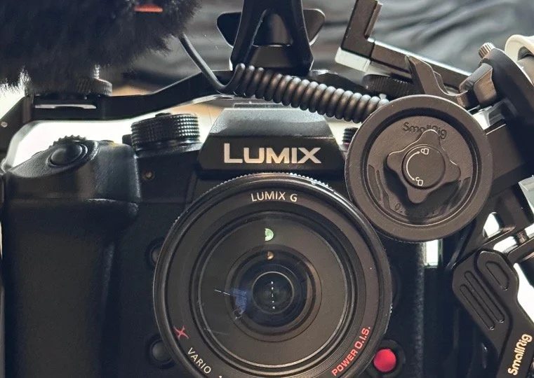 Close-up of a Panasonic Lumix DSLR camera with a large lens attached, mounted on a stabilizer rig with a coiled cable and monitor, used for professional videography or photography.