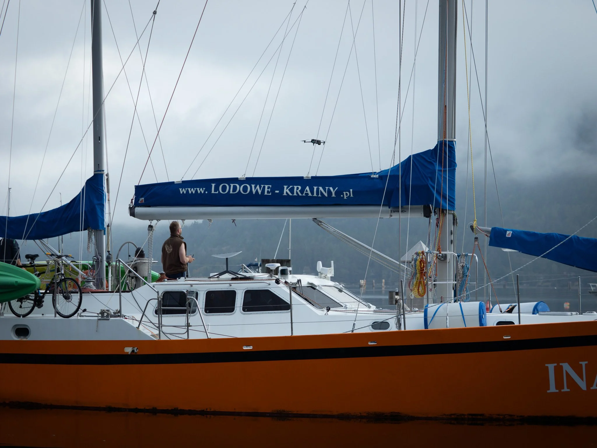 View of multiple sailboats docked at a marina under a cloudy sky, with one boat prominently displaying a website URL 'www.LODOWE-KRAINY.pl' on a blue sail cover. A person is seen on the boat, possibly preparing a drone flying nearby.