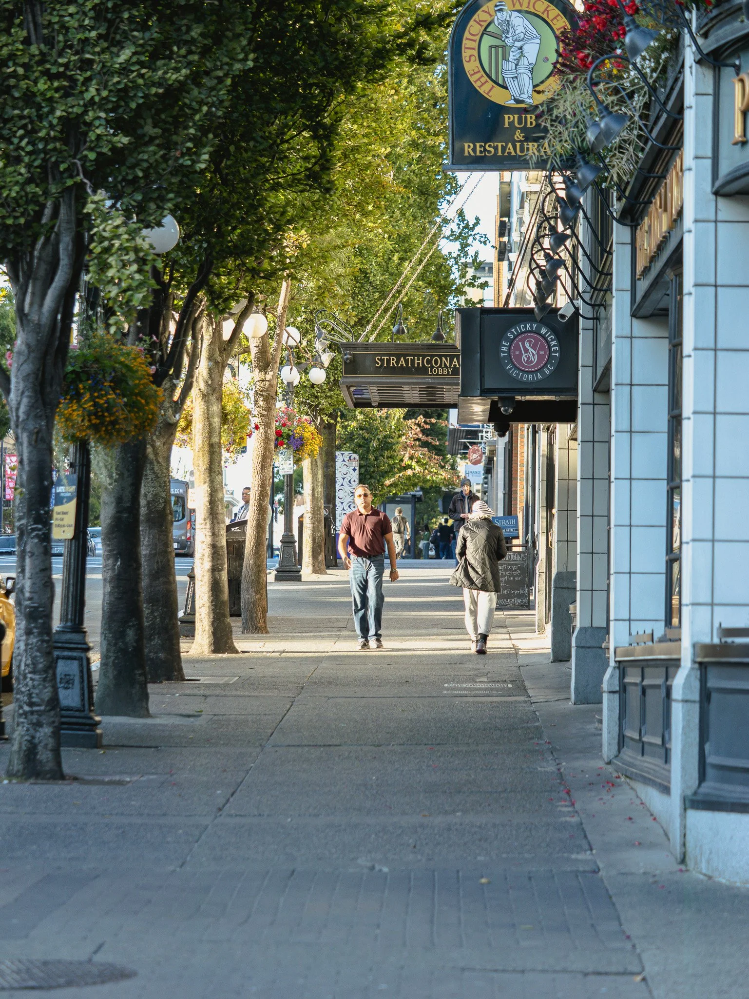Empty sidewalk in a downtown street with trees, overhead street lamps, and storefronts. Pedestrians are walking, and signs indicate businesses such as a pub and restaurant, and a lobby for The Strathcona in Victoria BC