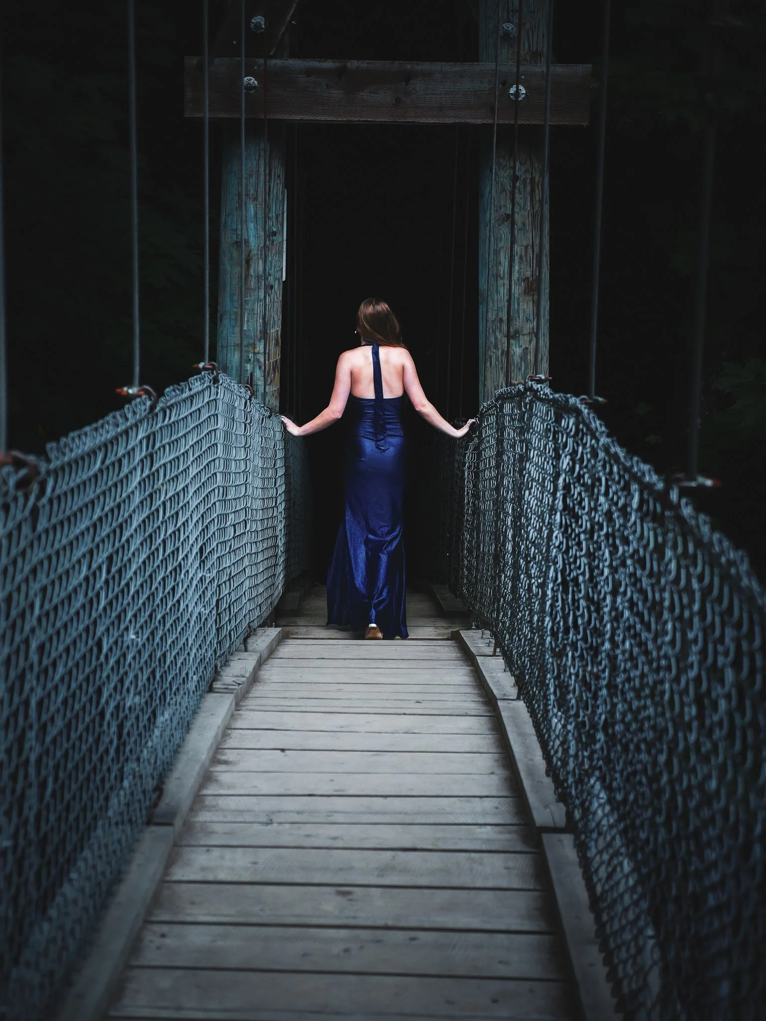Woman in a dark blue evening gown walking on a wooden suspension bridge with metal chain sides, surrounded by a dark, forested environment.