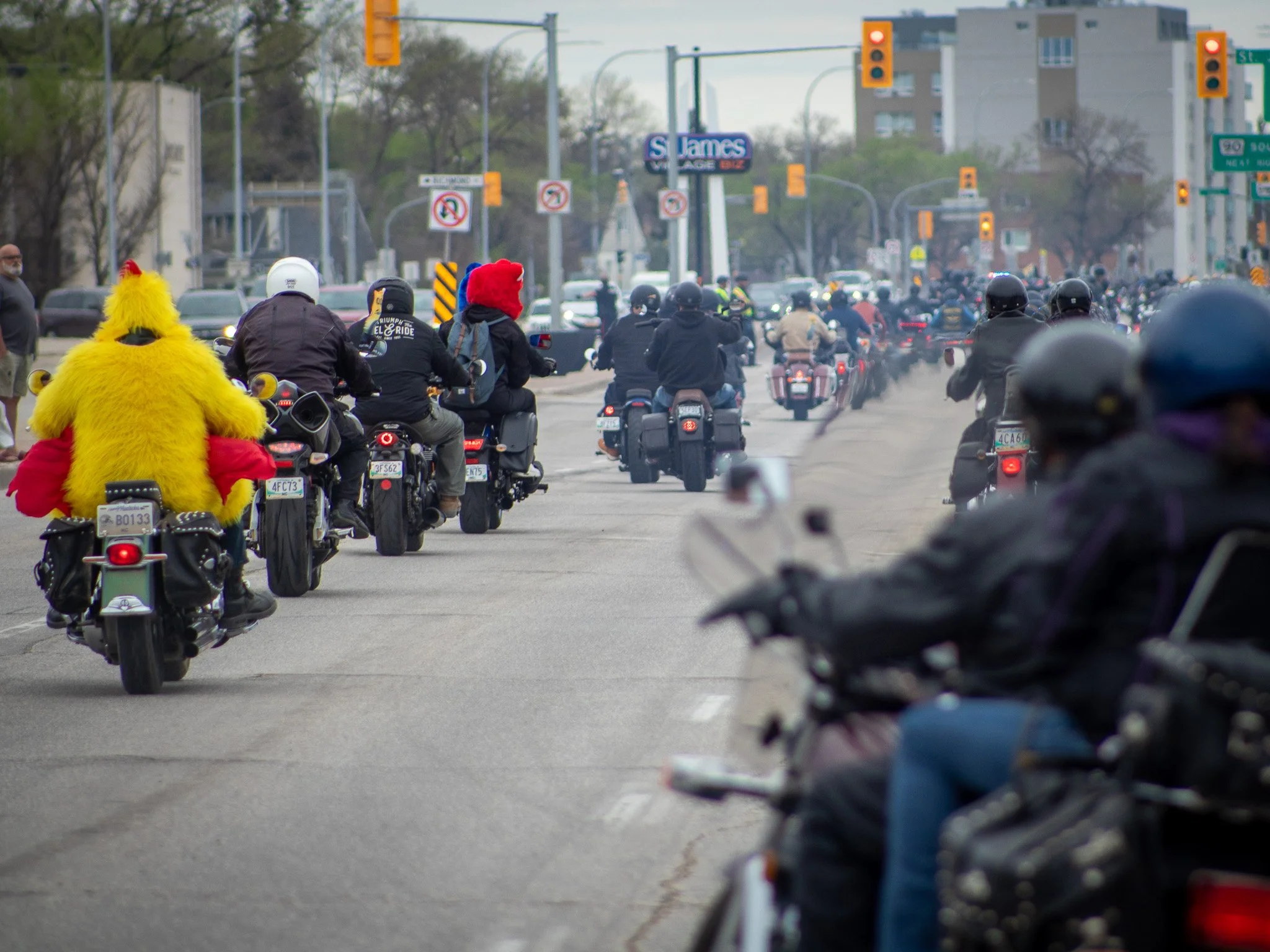 A group of motorcyclists riding together in an urban area with traffic lights and street signs visible. One rider is dressed as a chicken, wearing a bright yellow chicken costume with red accents. Other riders are wearing helmets and casual riding ge
