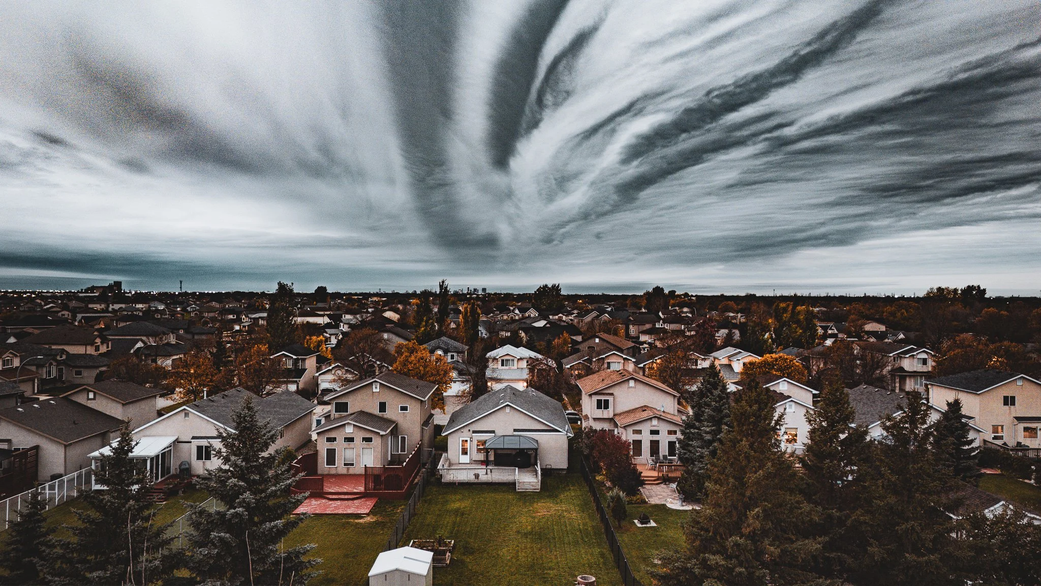 A suburban neighborhood with a clear view of houses, trees, and backyards under a dark, cloudy sky Winnipeg Manitoba.