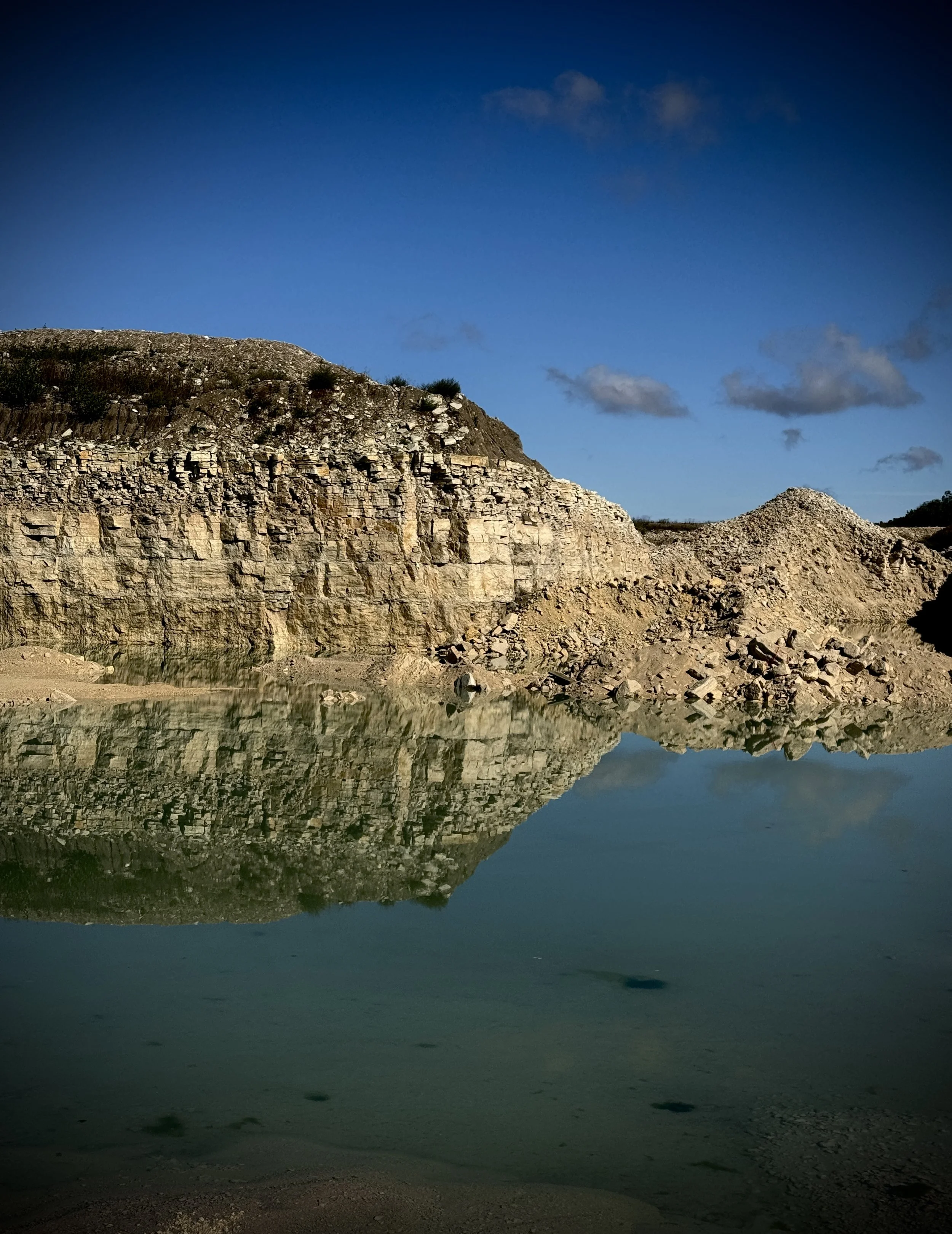 A peaceful landscape featuring rocky cliffs reflecting in calm water with a clear blue sky and scattered small clouds above.