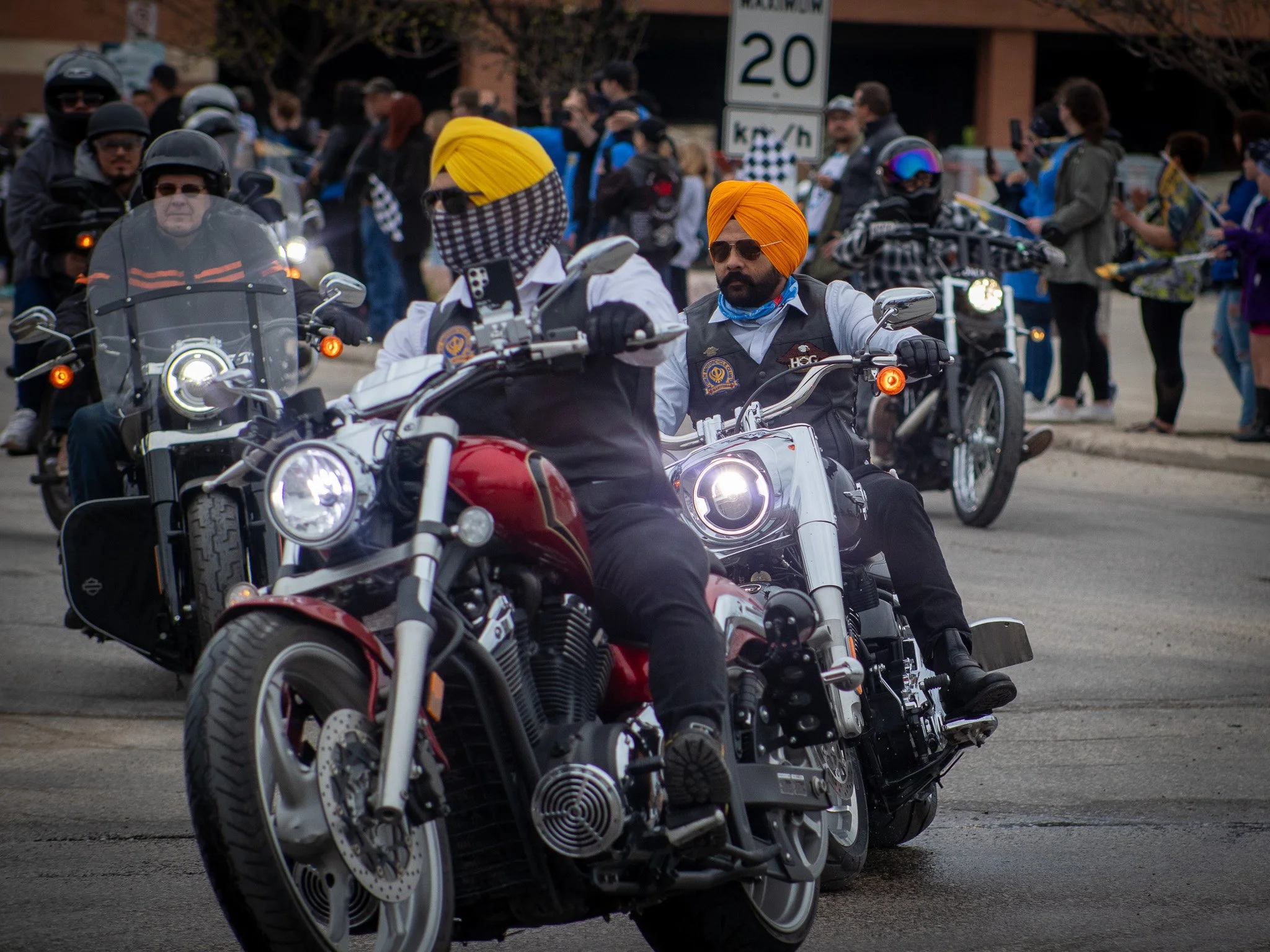 A group of motorcyclists, some wearing helmets and sunglasses, are gathering outdoors, with a sign indicating speed and other onlookers in the background ride for dad Winnipeg MB