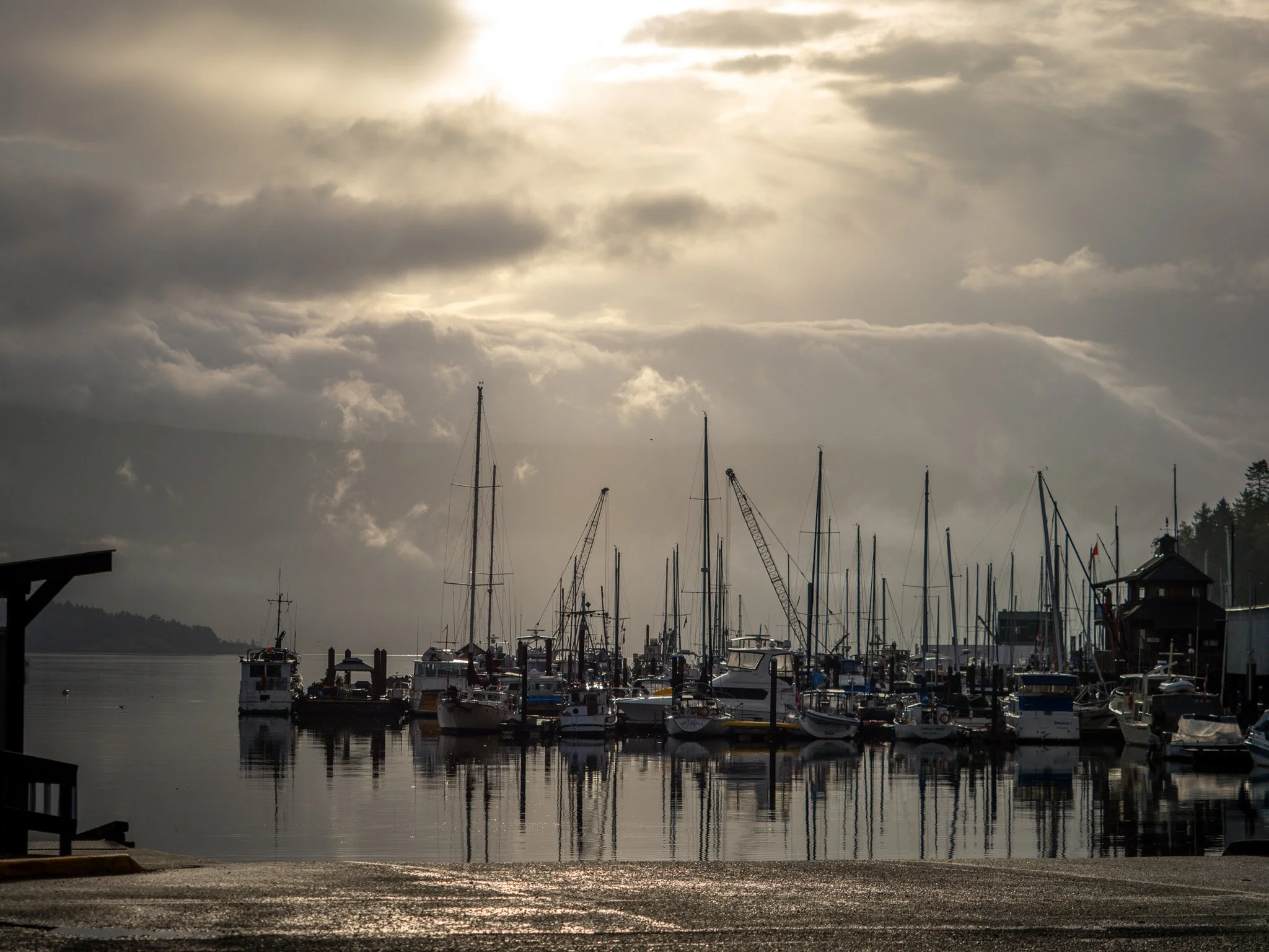 A marina with sailboats and yachts docked, calm water reflecting the boats, overcast sky with some sunlight breaking through clouds in Vancouver Island British Columbia Cowichan Bay 
