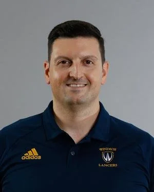Headshot of a man with short dark hair, smiling, wearing a navy blue polo shirt with Adidas logo and Wesleyan Lancers logo, against a plain gray background.