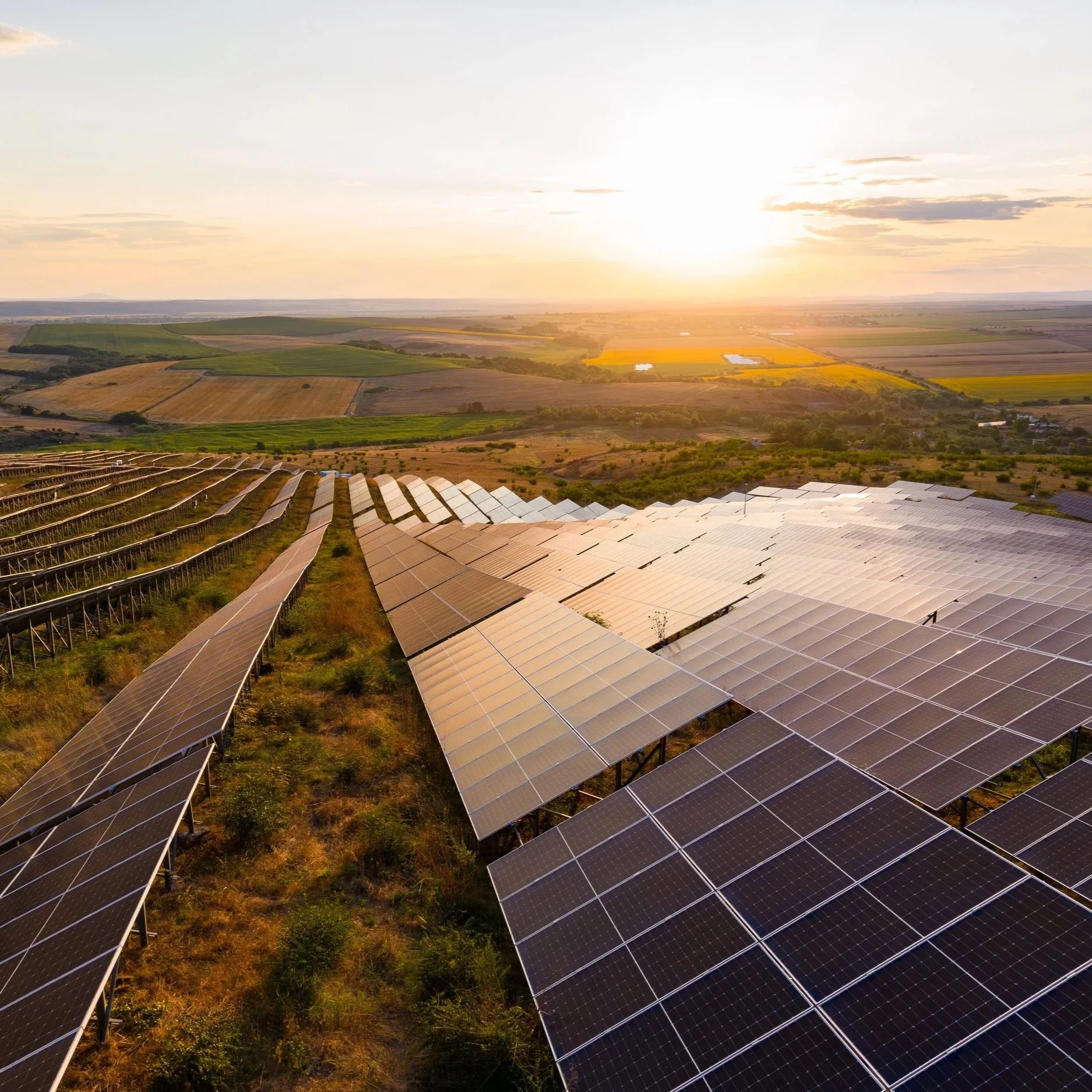 Solar panels installed on a hillside at sunset, overlooking agricultural fields and a distant horizon.