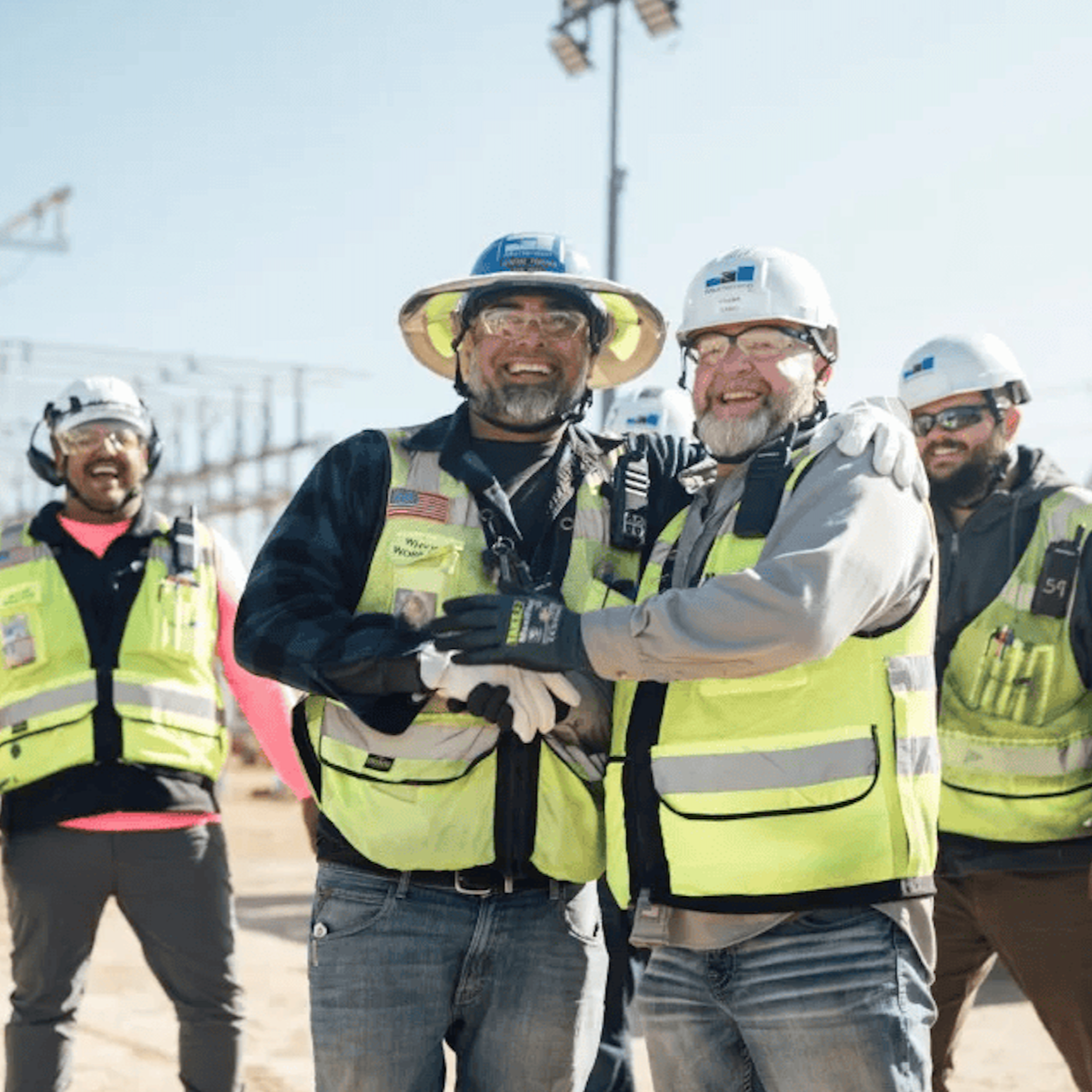 Group of construction workers in safety vests and helmets smiling and celebrating on a construction site with power lines and cranes in the background.