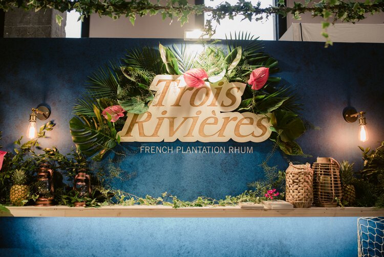 Enseigne "Trois Rivières" avec décoration tropicale, plantes vertes et fleurs sur un mur bleu, éclairée par des lampes murales.