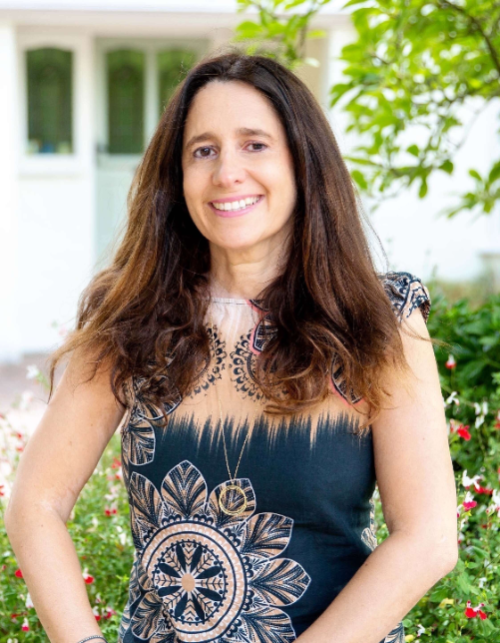 A portrait photograph of a smiling Amanda Stecker with long brown hair, wearing a dark patterned dress and posing outdoors with green bushes and a building in the background