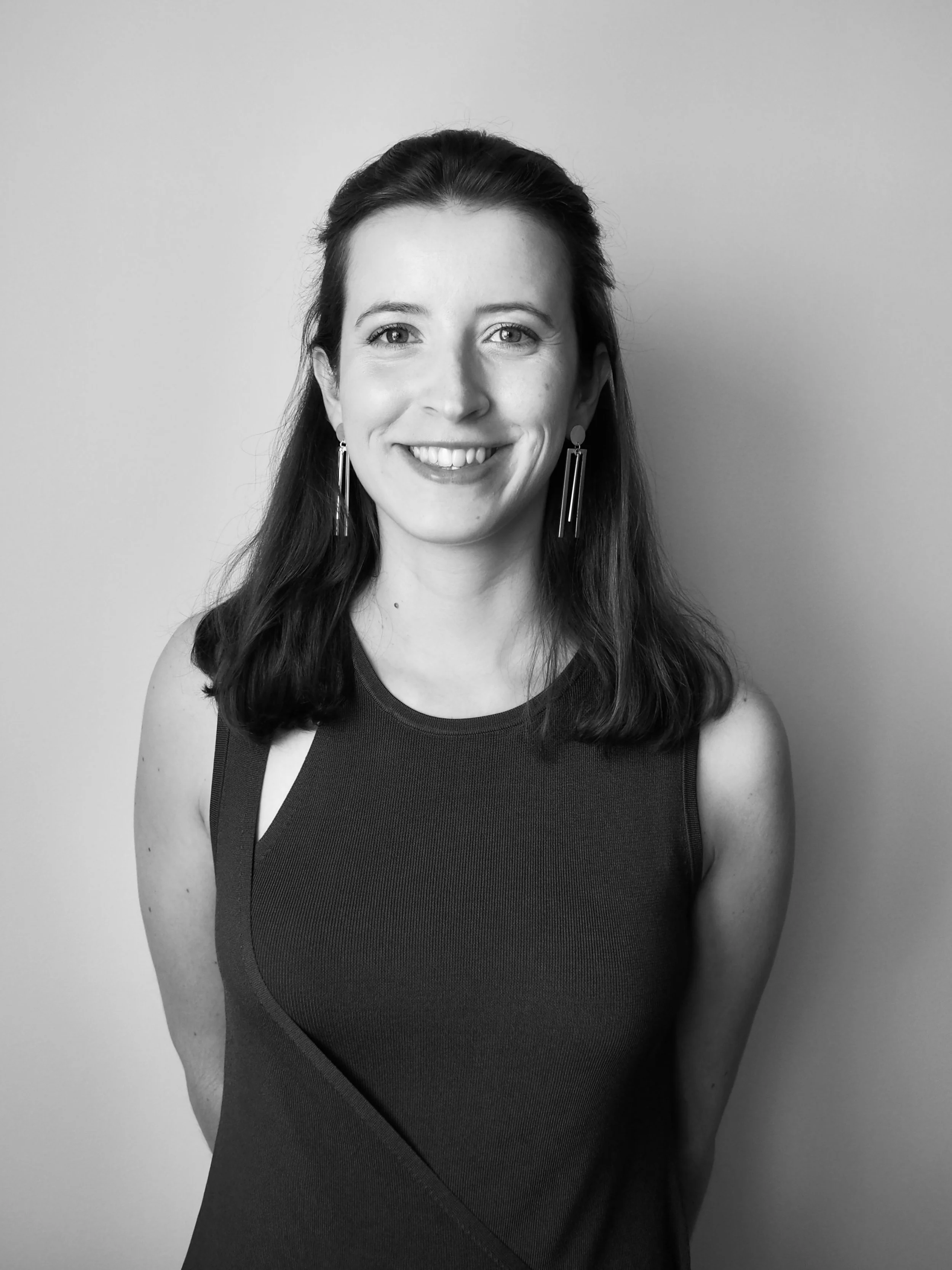 Retrato em preto e branco de uma mulher sorridente, com cabelo escuro médio, vestindo uma blusa sem mangas e brincos longos, frente a uma parede clara.