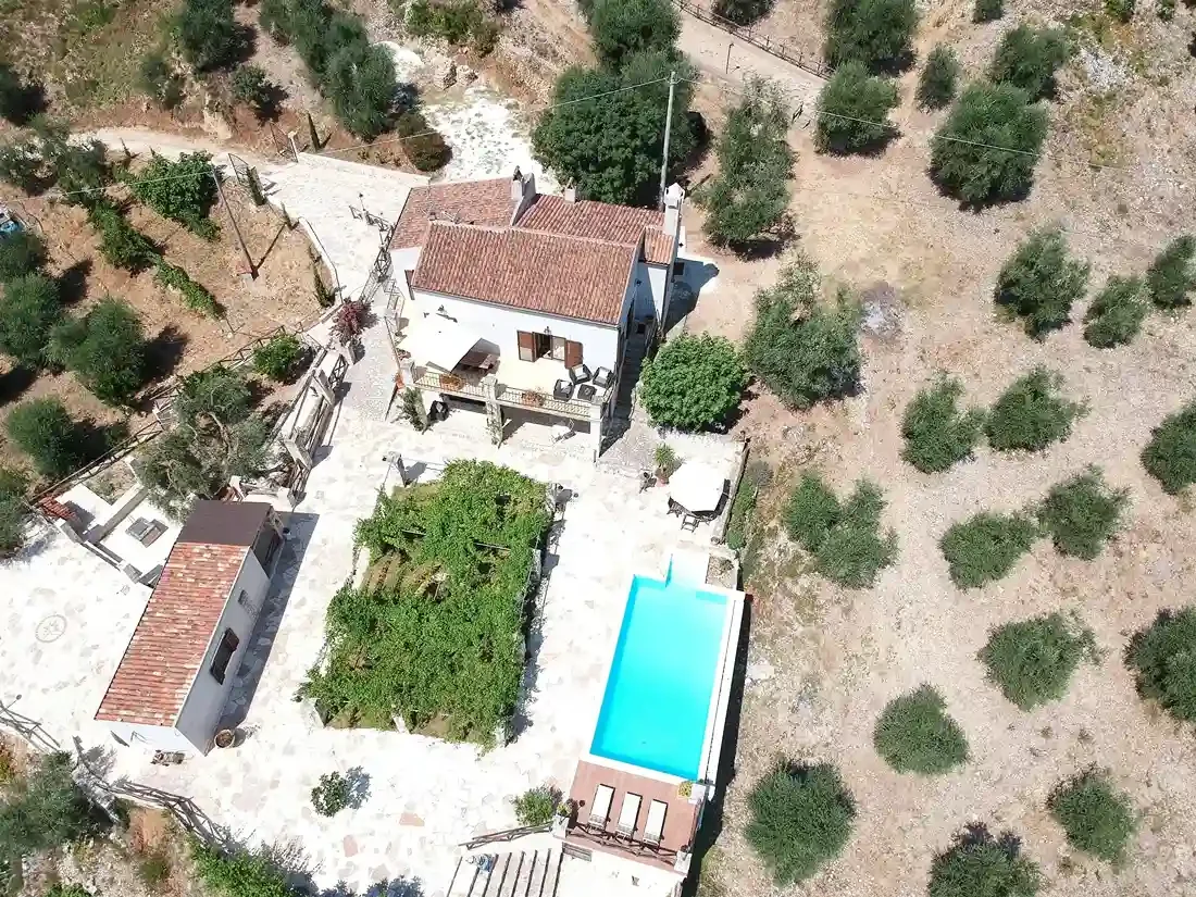 Aerial view of a house with a tiled roof, surrounded by a patio with outdoor furniture, trees, and a swimming pool, with fruit trees and dry land around.