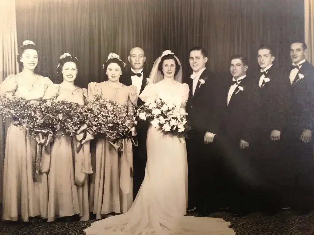 Black and white photo of a wedding party with six women in bridesmaid dresses holding large bouquets, a bride in a white gown with a veil and bouquet, and five men in tuxedos with bow ties and boutonnières, standing against a curtain backdrop.