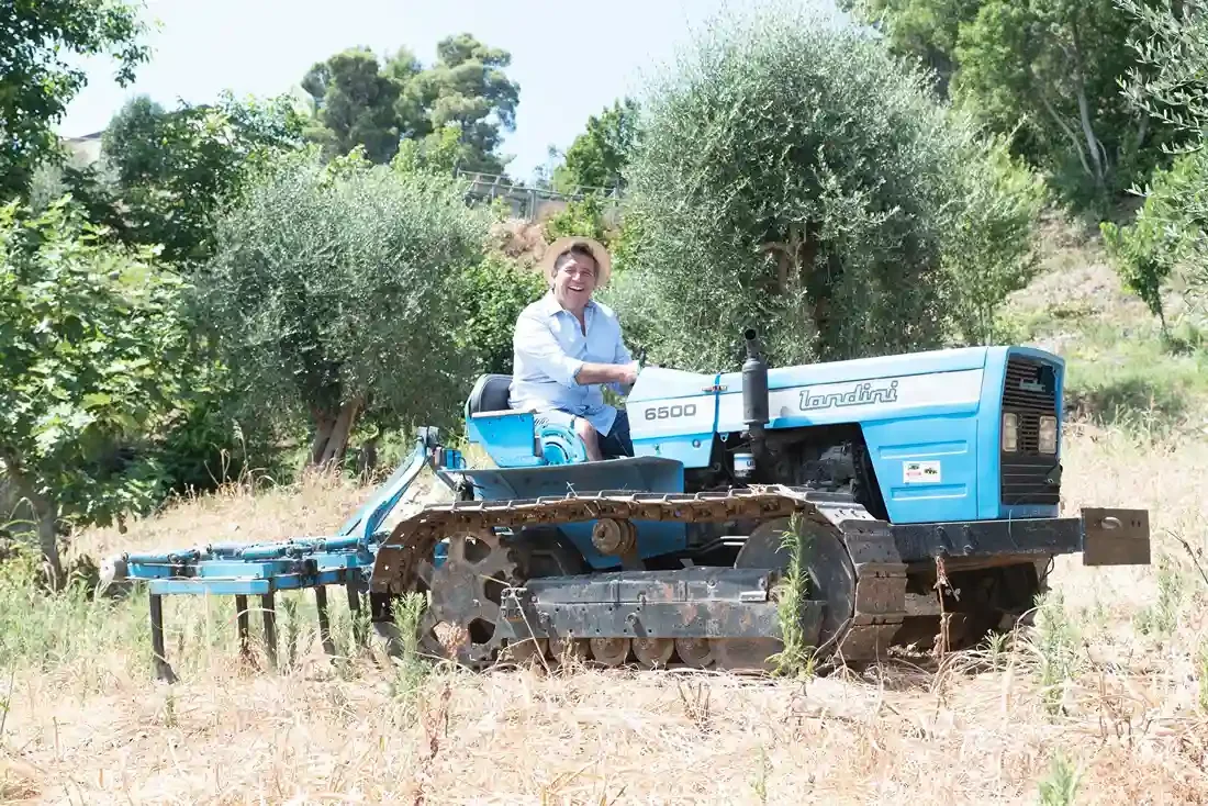 A smiling man sitting on a blue tractor in a farm or orchard surrounded by green trees and bushes during daytime.