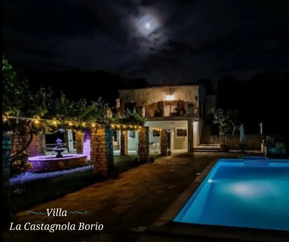 Nighttime view of Villa La Castagnola Borio showing an illuminated swimming pool, string lights, a fountain, and a terrace under a cloudy moonlit sky.