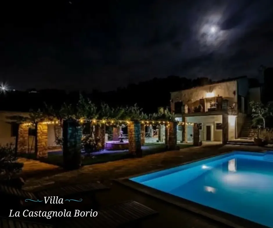 Night shot of a villa named La Castagnola Borio with a lit swimming pool and outdoor area decorated with string lights, under a cloudy sky with the moon.