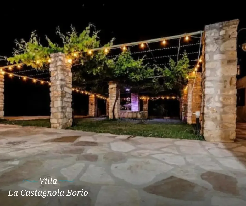 Night scene of a stone villa patio decorated with hanging string lights, surrounded by trees and stone pillars at La Castagnola Borio.