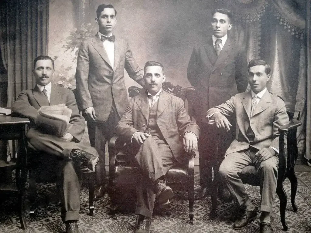 Black and white photograph of five men in vintage suits posed in a formal setting, with two seated and three standing, in front of a painted backdrop and patterned carpet.