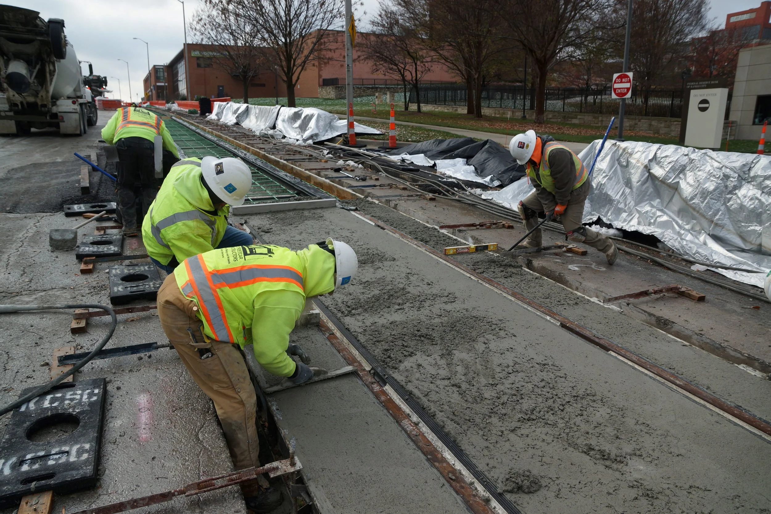 Image of four crew members spreading out freshly poured concrete for newly built track. Crews are bundled up with hats, gloves and warm coats. Blankets are lined along the curb in preparation of covering the concrete.