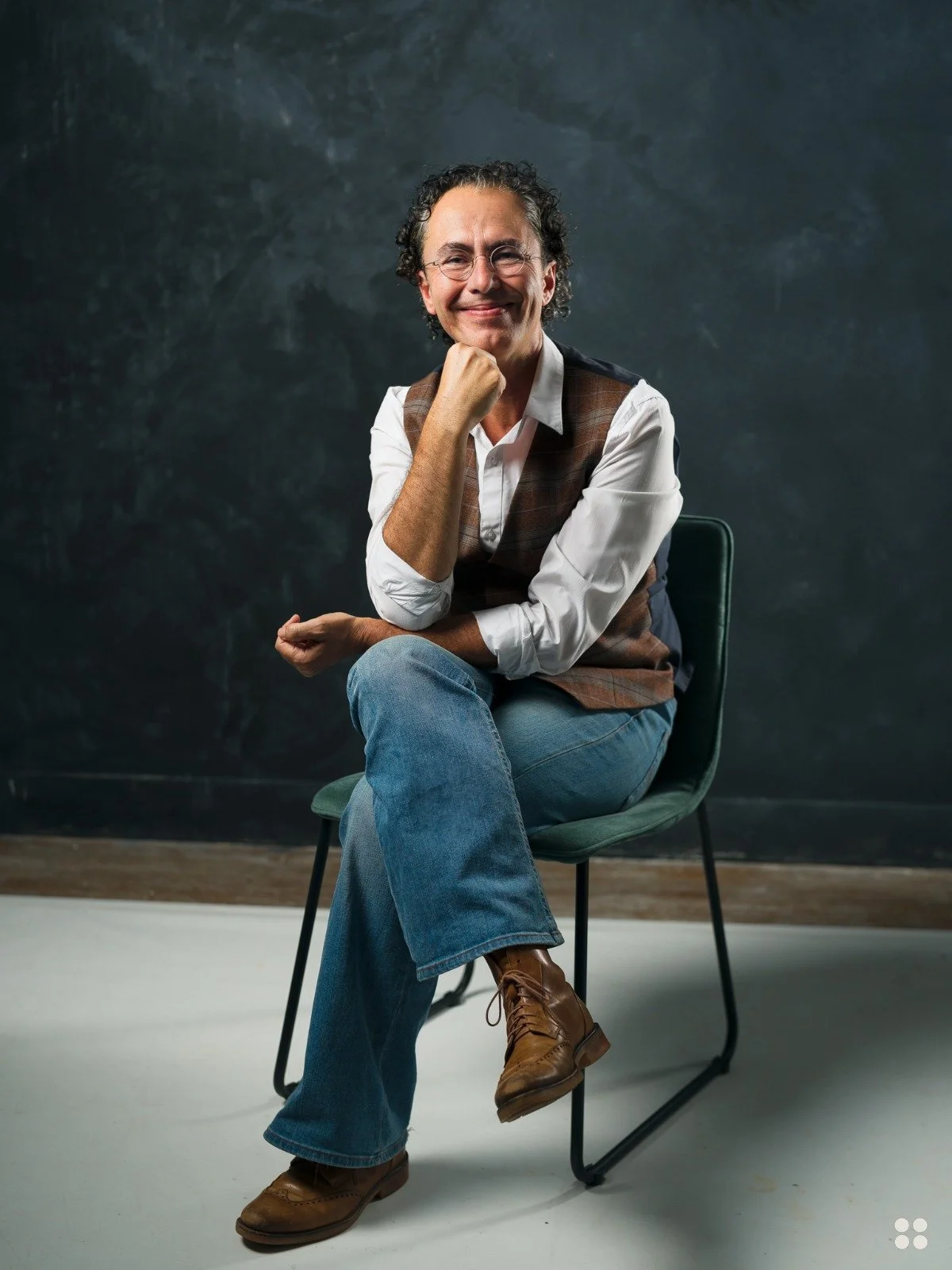 A man with curly hair and glasses, wearing a white shirt, vest, and jeans, sitting on a chair with one leg crossed over the other, smiling at the camera with a dark background.