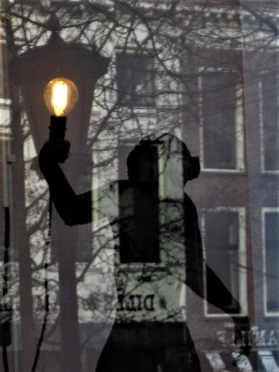 Reflection of a person holding a lit street lamp and a camera, with trees and buildings visible in the background.