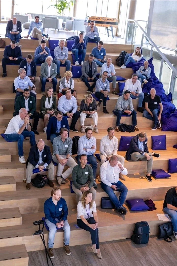 Group of people attending a conference or seminar, seated on wooden tiered seating with purple cushions, in a bright modern space with large windows.