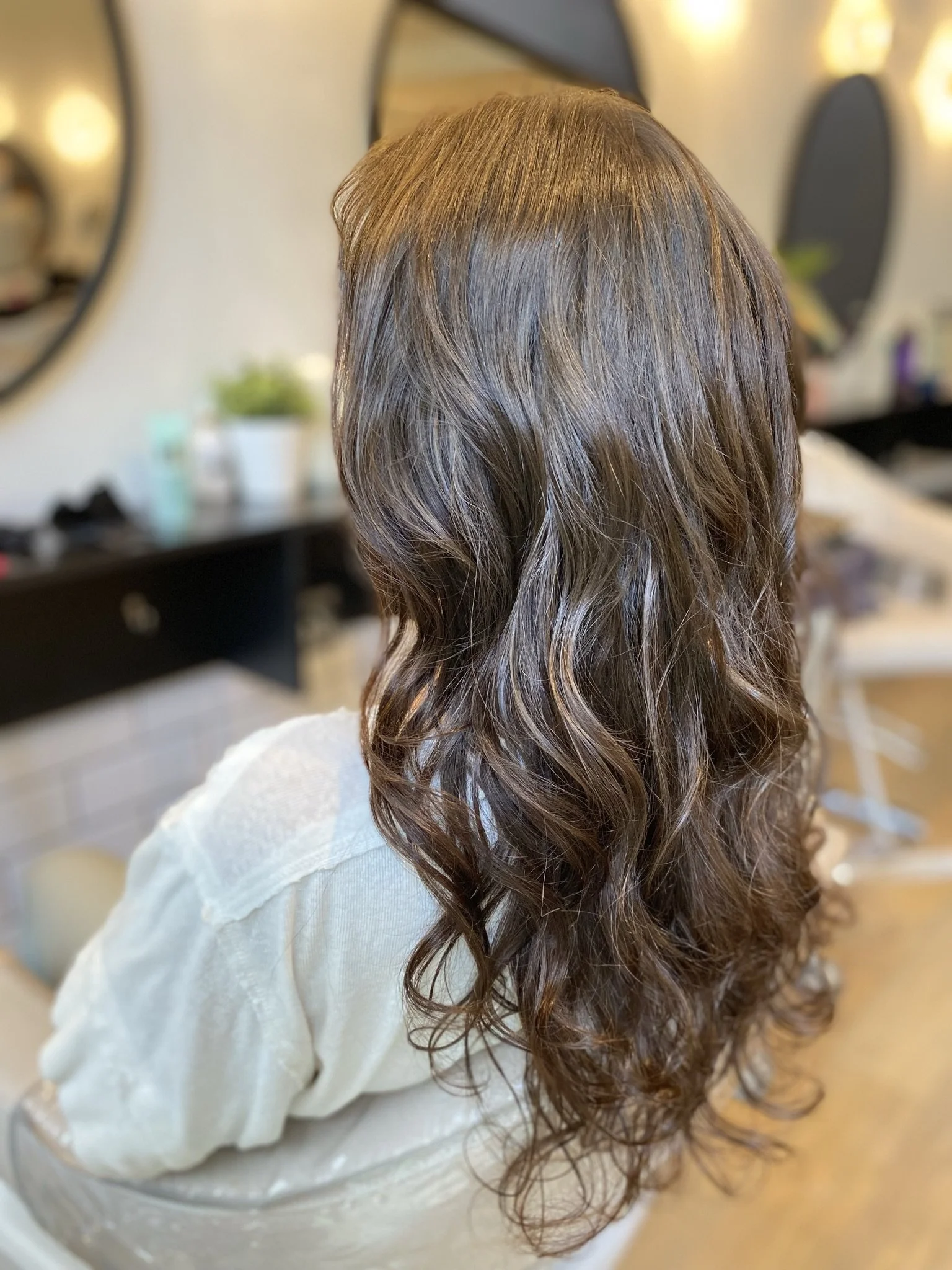 Back view of a woman with long, wavy brown hair at a salon.