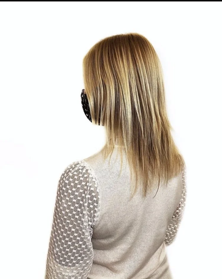 A woman with shoulder-length blonde hair wearing a gray sweater with textured sleeves and a black face mask, facing away from the camera against a plain white background.