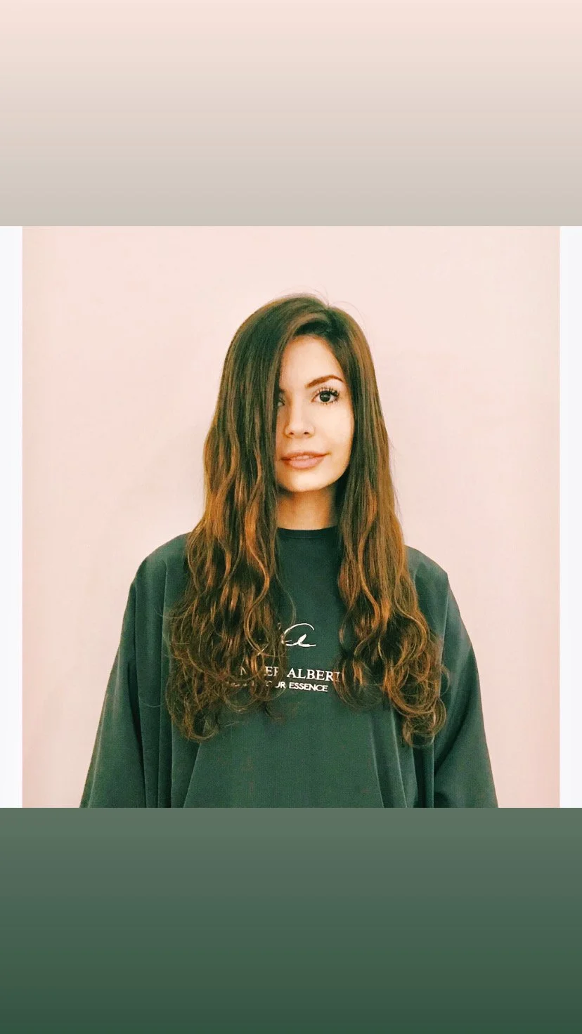 A woman with long wavy brown hair, wearing a dark sweatshirt, looking at the camera with a neutral expression, standing against a light-colored wall.