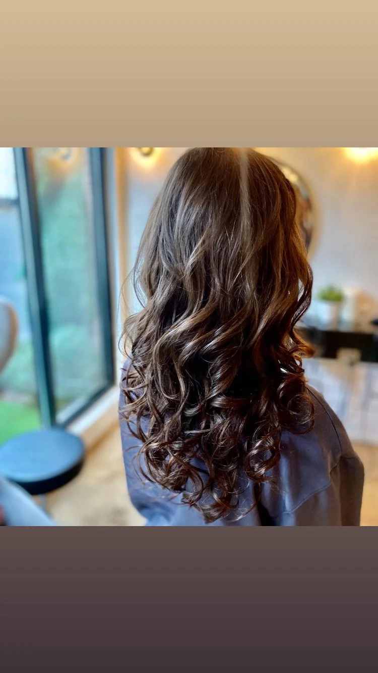 A woman with long, dark, curly hair standing indoors near large windows, with a blurred background of a salon or modern room.