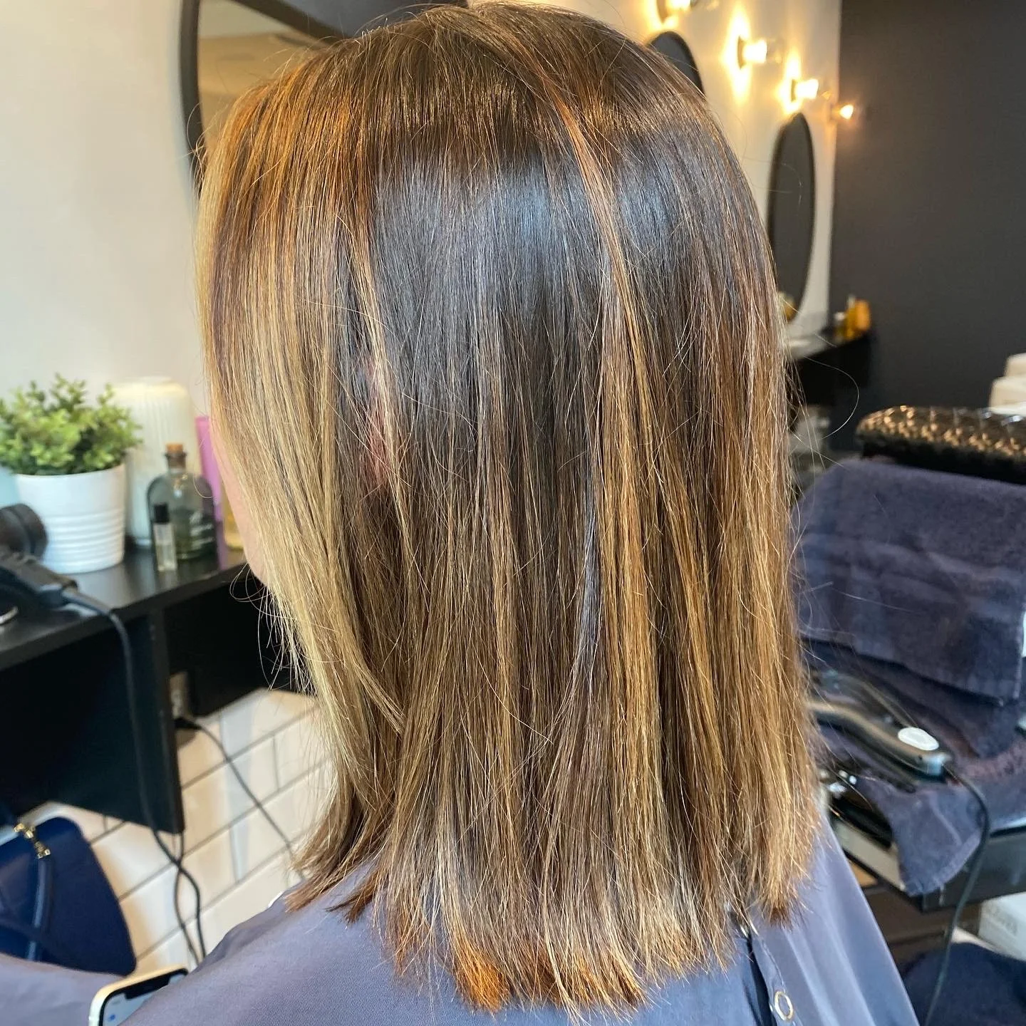 The image shows a person with shoulder-length brown hair with highlights, sitting in a salon chair, with a mirror and salon tools in the background.