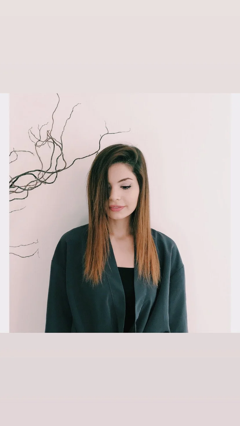 A young woman with long brown hair, wearing a black top and oversized black jacket, standing against a white wall decorated with black, twisting branches.