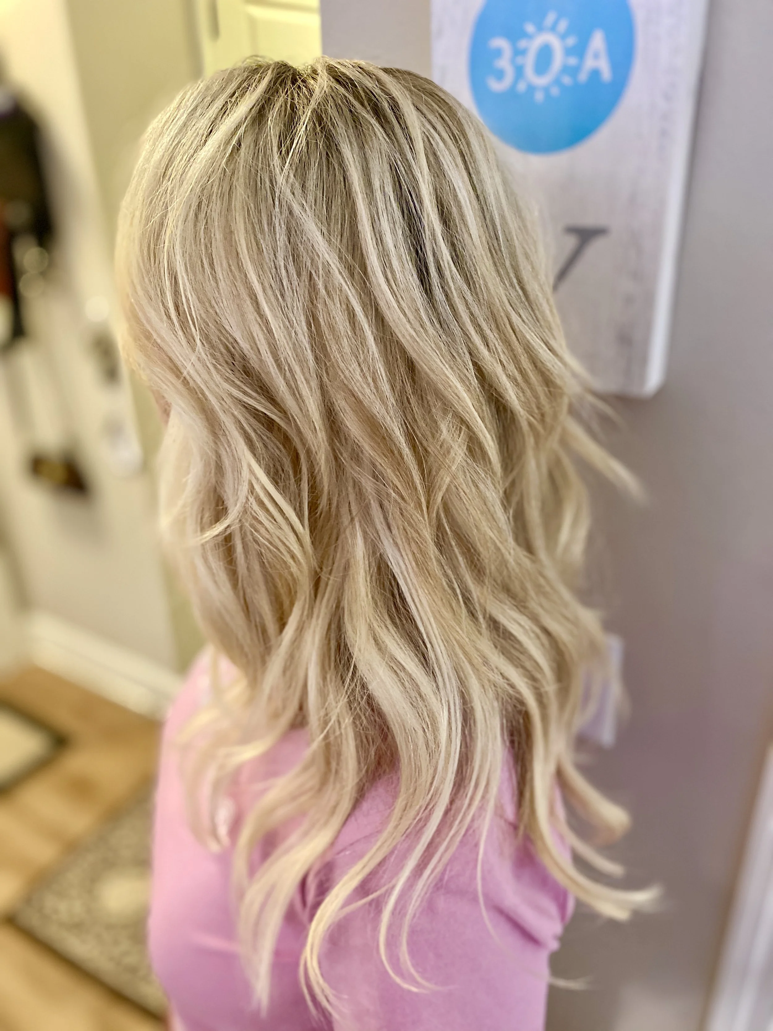 A woman with long, wavy blonde hair, wearing a pink top, standing indoors next to a wall with a blue and white sign.