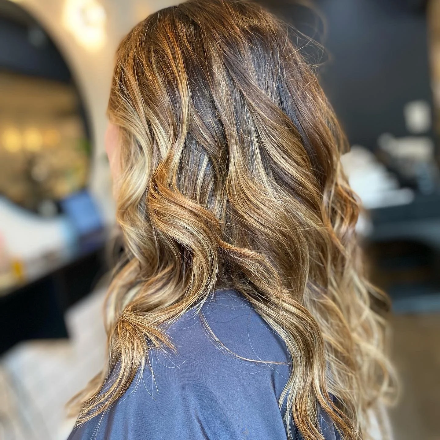 Close-up of a woman's wavy, shoulder-length hair with highlights, seen from the back in a salon.