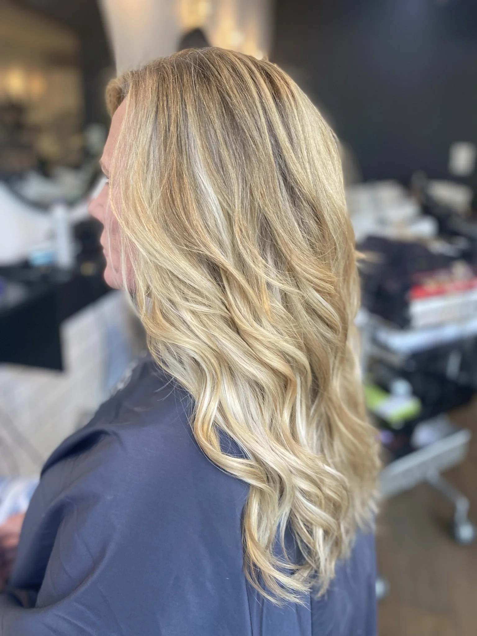 Side view of a woman with long, wavy blonde hair sitting in a salon chair, with salon tools and products in the background.