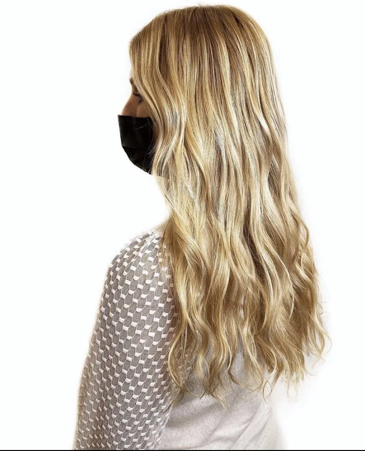 Side view of a woman with long, wavy blonde hair wearing a black face mask and a white textured sweater, standing against a plain white background.