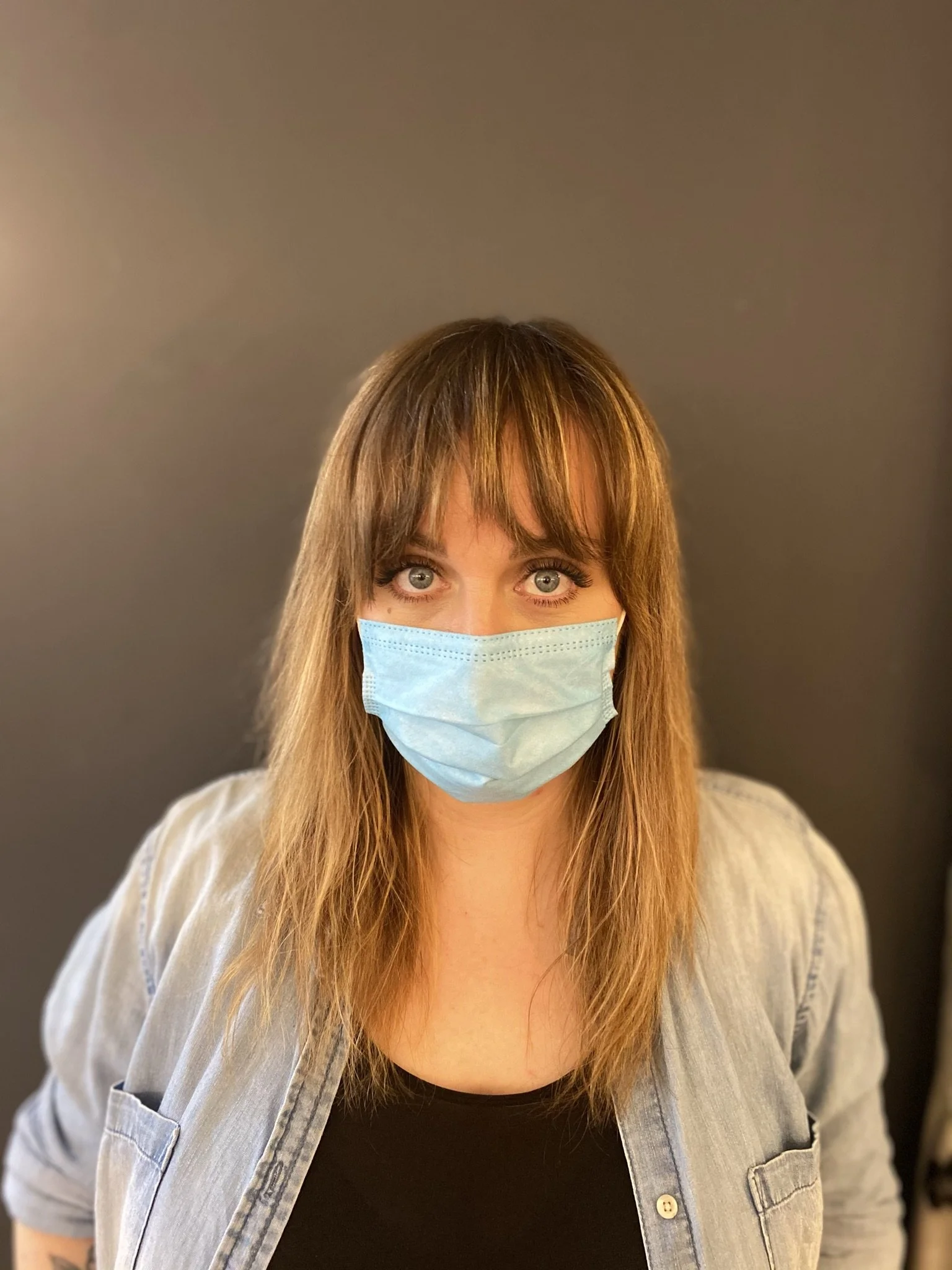 A woman with long brown hair and blue eyes wearing a light blue face mask, a black top, and a light gray denim shirt, standing in front of a dark gray background.