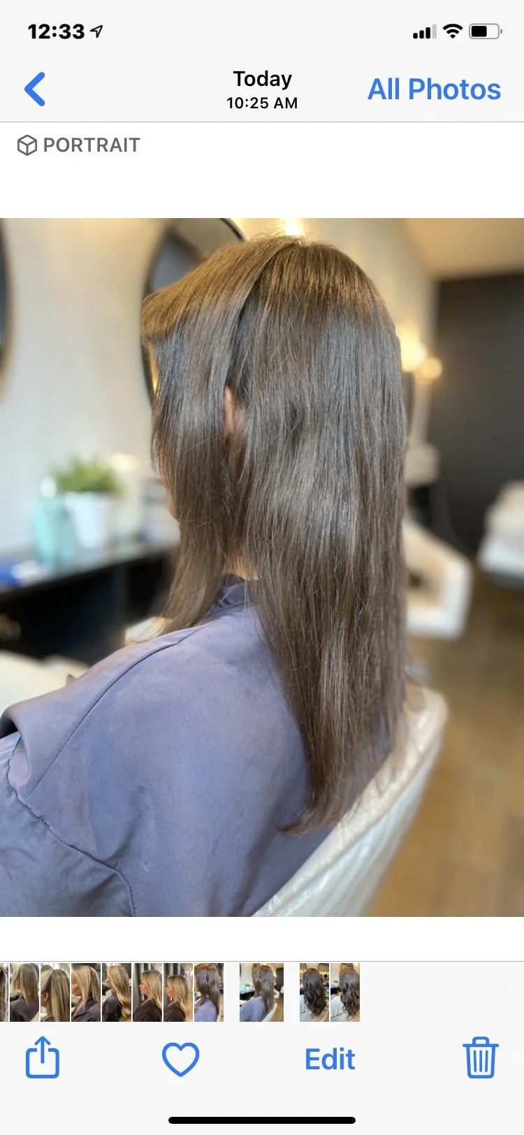 Side view of a woman with straight, shoulder-length brown hair, seated in a salon chair with a dark wall and a plant in the background.