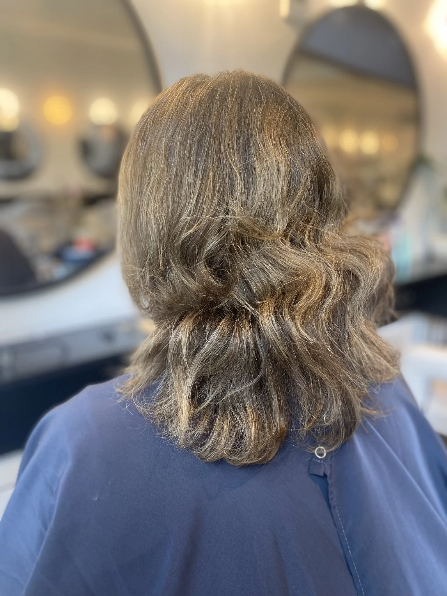 Back of a woman with shoulder-length wavy brown hair, sitting in a salon chair, with mirrors and salon decor visible in the background.