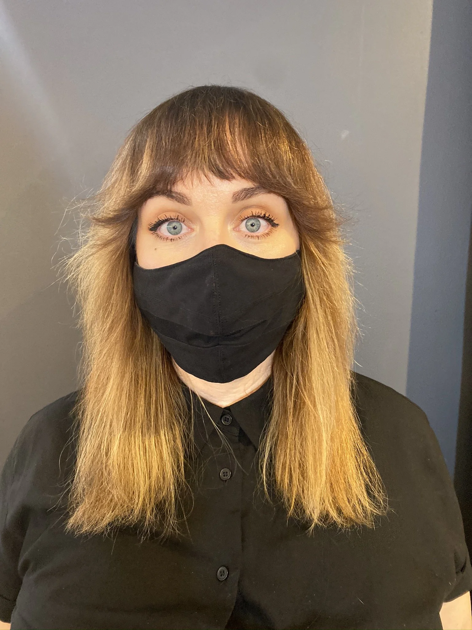 A woman with long reddish-brown hair and blue eyes wearing a black face mask and black shirt, standing against a plain gray wall.
