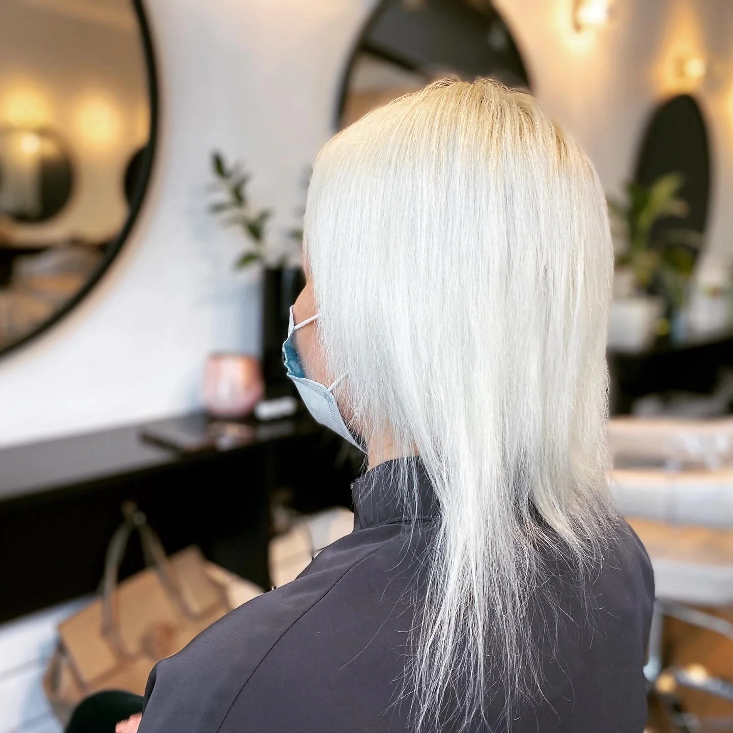 Side view of a woman with long, straight, platinum blonde hair wearing a protective face mask, sitting in a salon with mirrors and plants in the background.