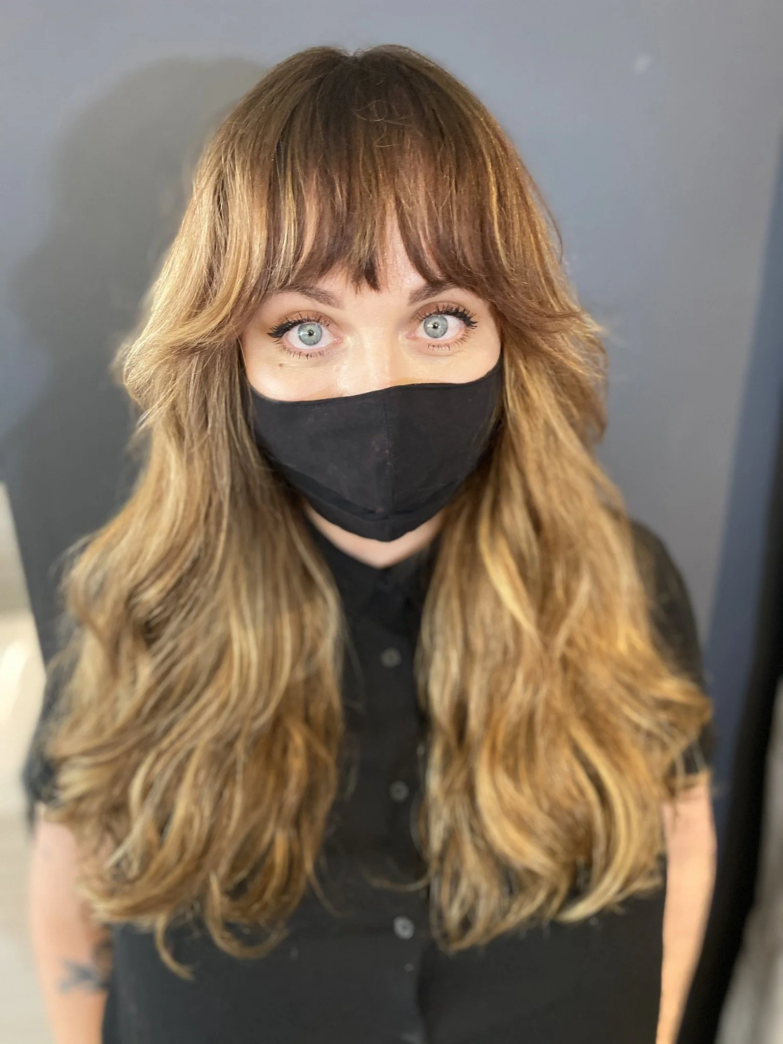 A woman with long, wavy, light brown hair with blonde highlights wearing a black face mask and black shirt, standing against a gray wall.