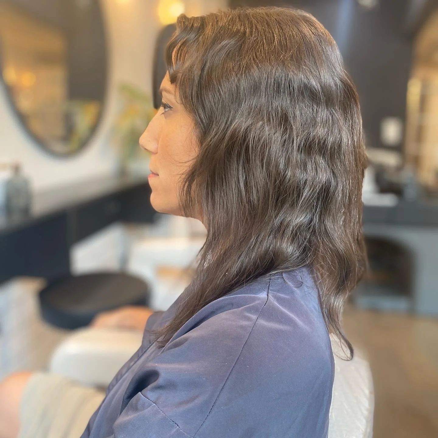 Side profile of a woman with wavy dark brown hair sitting in a salon chair.