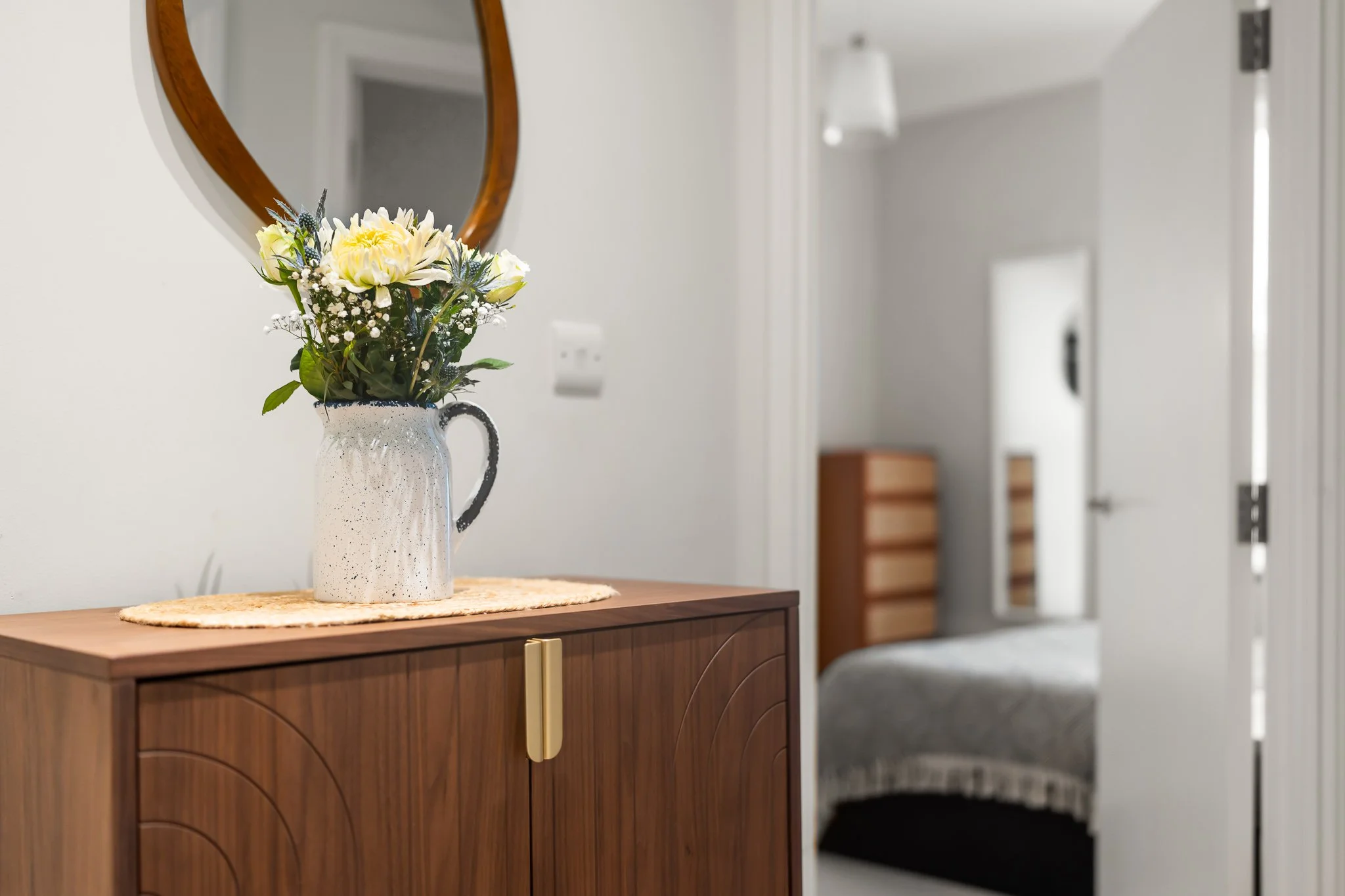 A room interior with a wooden cabinet topped with a ceramic pitcher holding a flower arrangement of white and yellow flowers. A round mirror is on the wall above the cabinet, and there is a bed in the background with a window and a brown dresser.