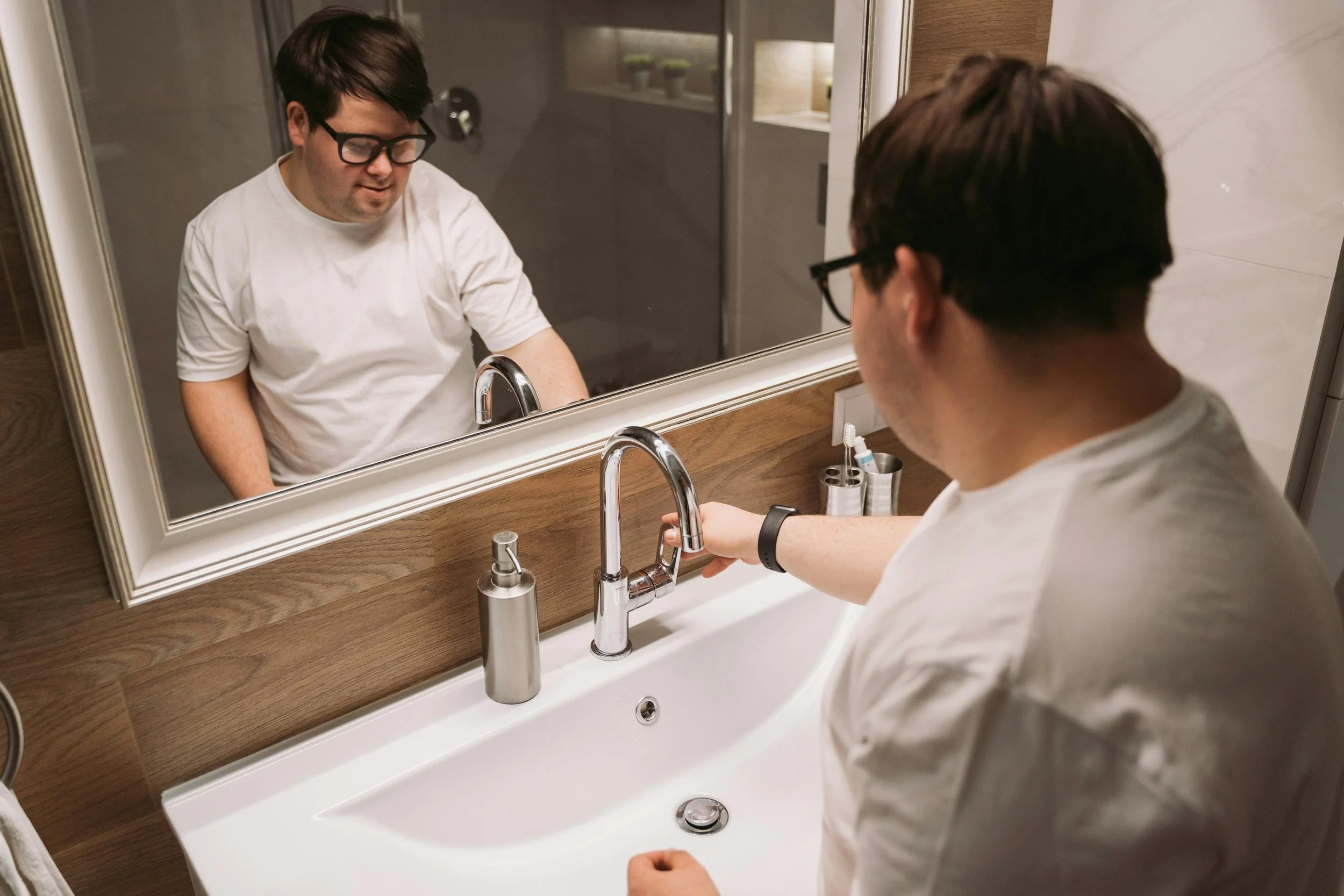A man with glasses and a white t-shirt stands at a bathroom sink, looking into the mirror and reaching for the faucet. The bathroom has a wooden panel behind the sink, a soap dispenser, toothbrush holder, and a reflection of shelves with green plants.